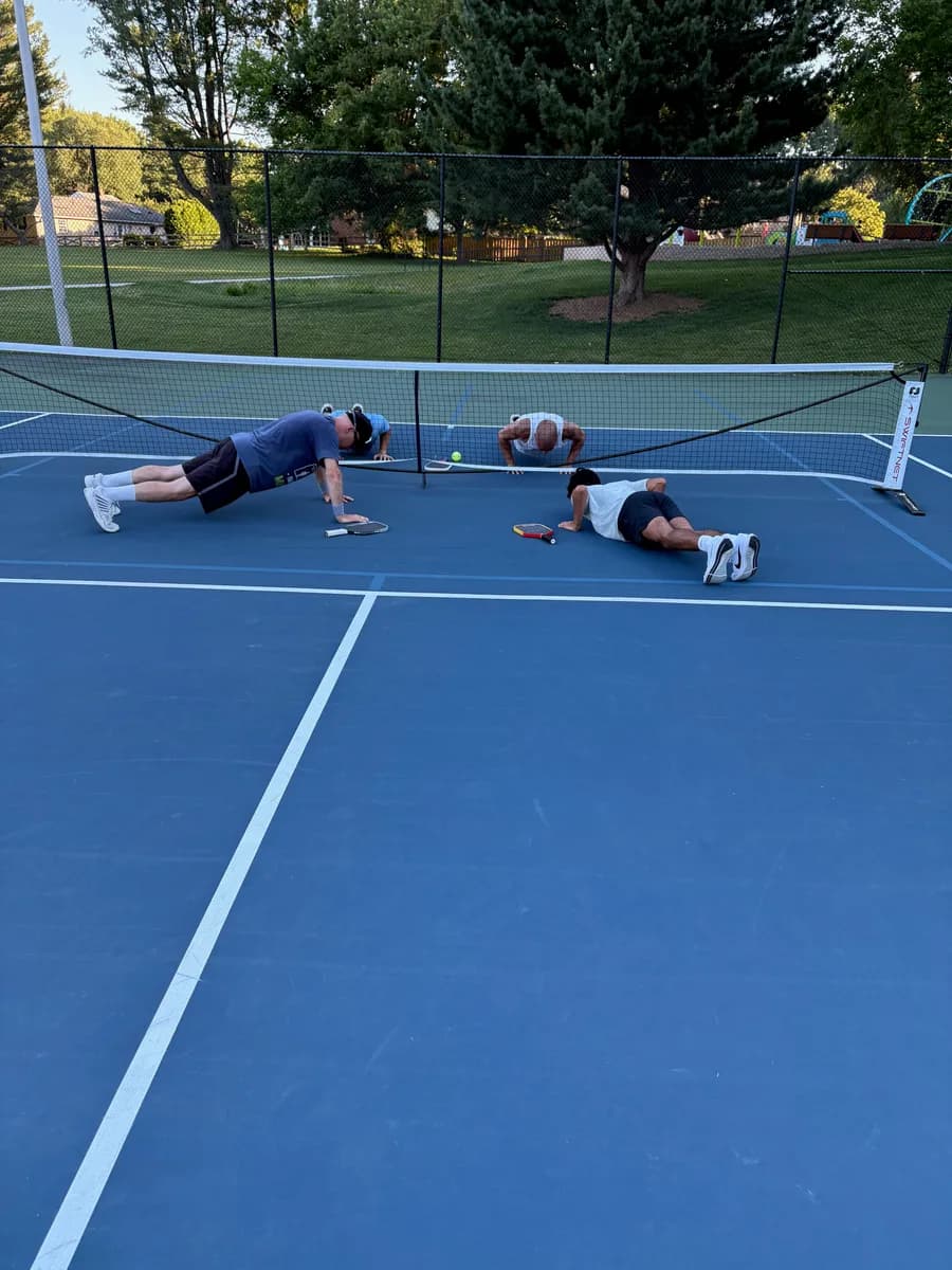 Four adult pickleball players doing pushups beside an outdoor net
